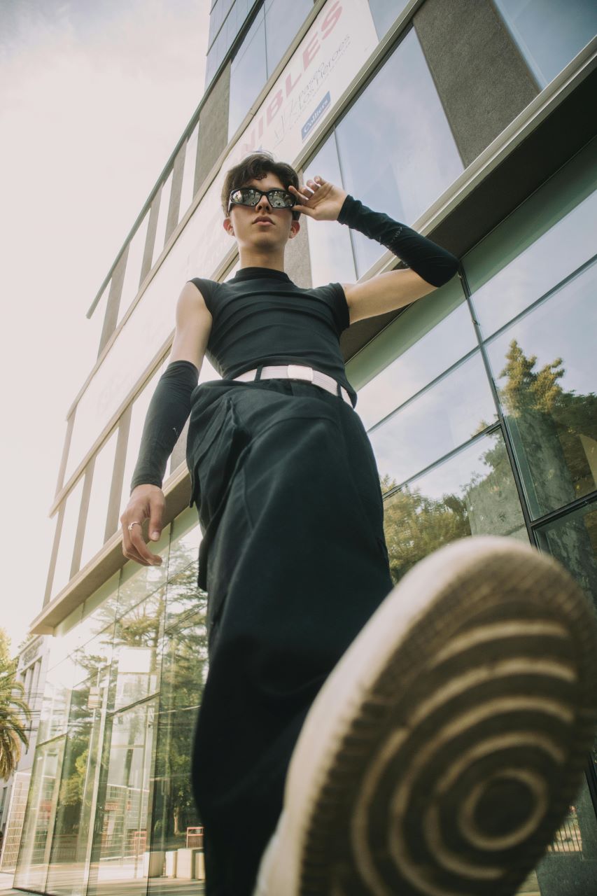 A young man stepping forward, camera angle looking up from the shoe coming towards it. He's wearing black pants, a black sleeveless top, a white belt, black arm wraps around his forearms, and reflective sunglasses