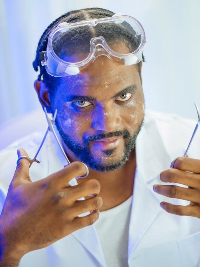 A man in a lab coat with goggles holding medical tools and looking menacingly at the camera, a blue light on half his face