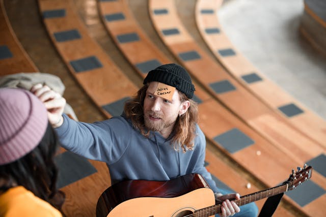 A man holding a guitar has a note on his forehead that says Julius Caesar
