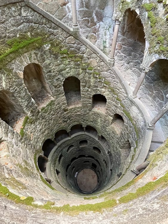 A circular well surrounded by gray stone stairs. Arched windows look into the well. Moss grows on the walls.