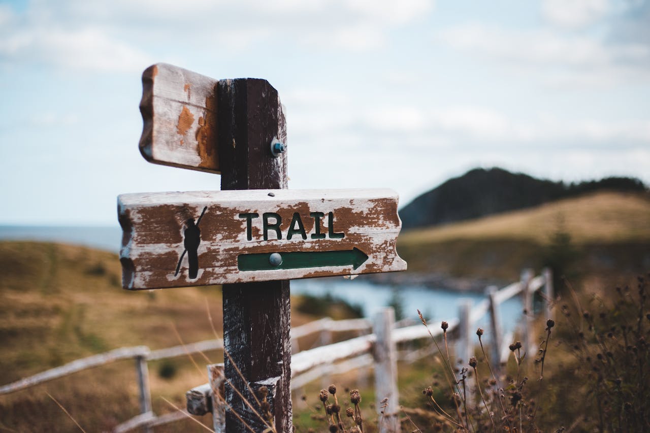 Backdrop of rolling countryside with pale blue sky. A post-and-rail fence crosses the lower half and a weathered post with directional signs takes foreground. On one brown plank, a black arrow points right and says "Trail."