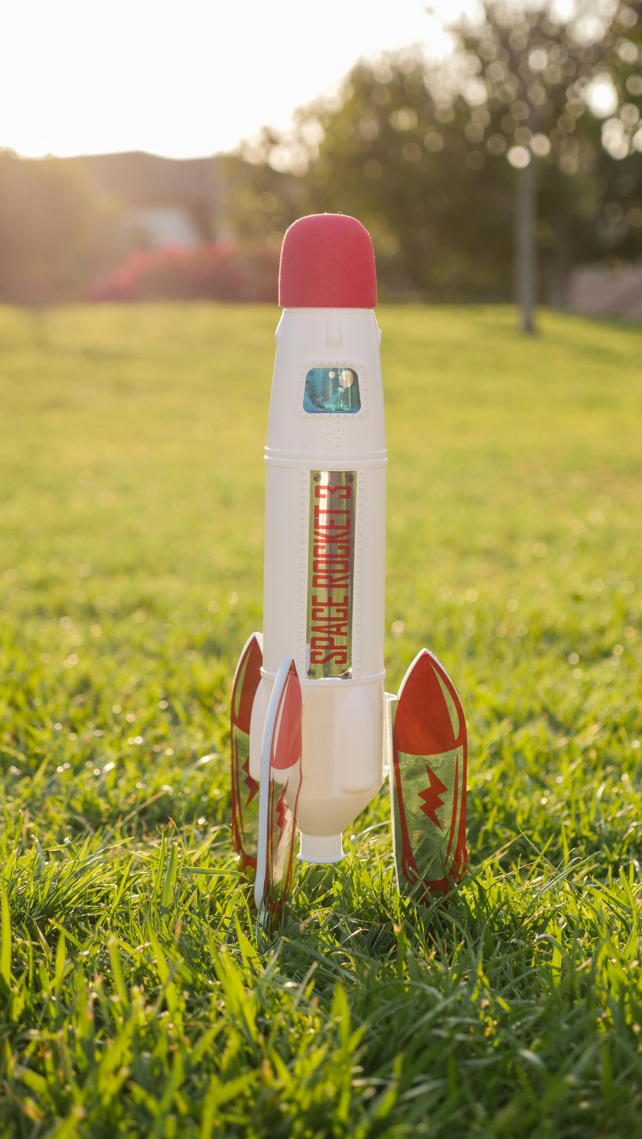 A white bottle rocket with red accents and red and gold engines supporting it sits on a sunny, grassy field